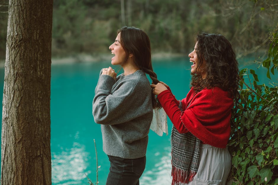 Two women enjoying a playful moment by a turquoise lake, styling hair amidst nature.