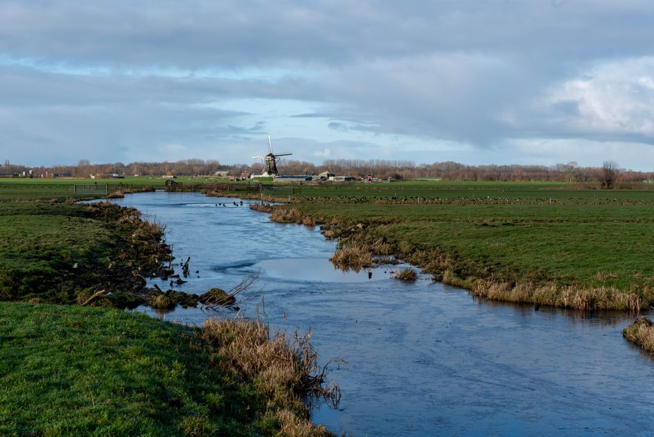 Idyllic rural scene featuring a serene river and traditional Dutch windmill under a cloudy sky.