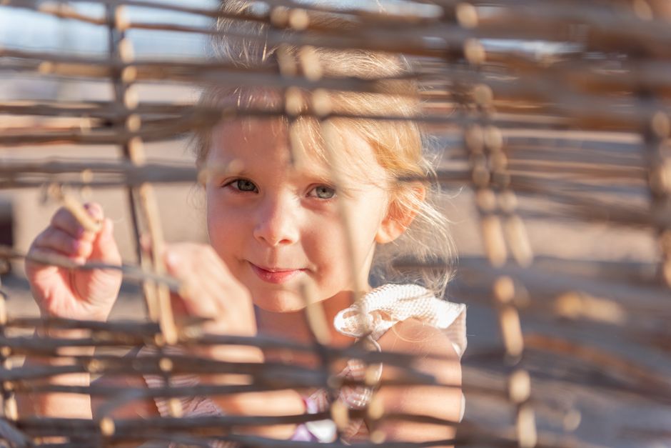 Cute little girl standing on sandy beach and looking at camera through straw fence during summer holidays