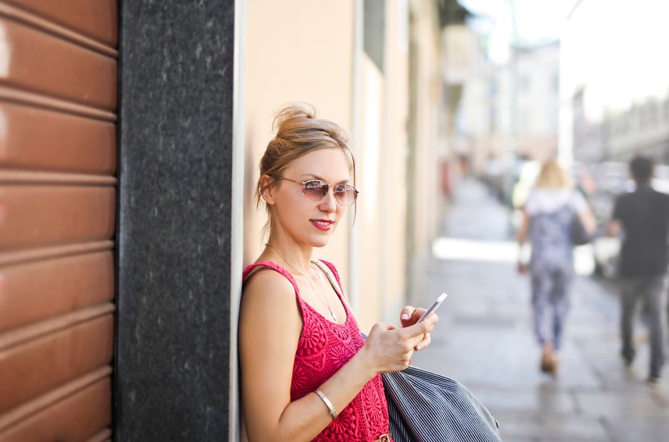 Fashionable woman wearing sunglasses and holding phone, enjoying sunny urban street.