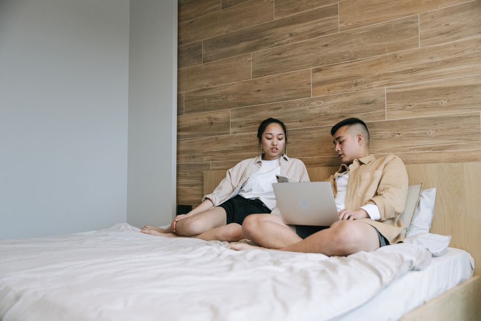 Asian man and woman sitting on bed using a laptop for work or leisure in a modern cozy room.