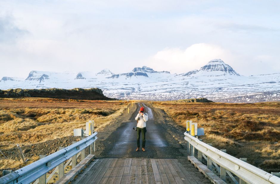 A photographer stands on a road capturing snow-capped mountains with a camera.
