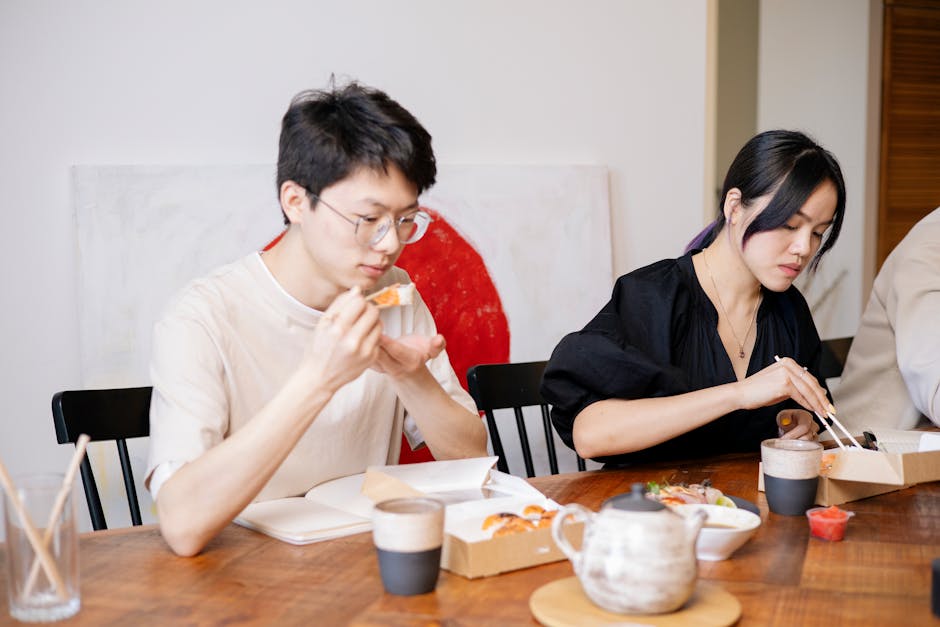 An Asian couple sharing a meal with chopsticks at a wooden table indoors, enjoying casual dining.