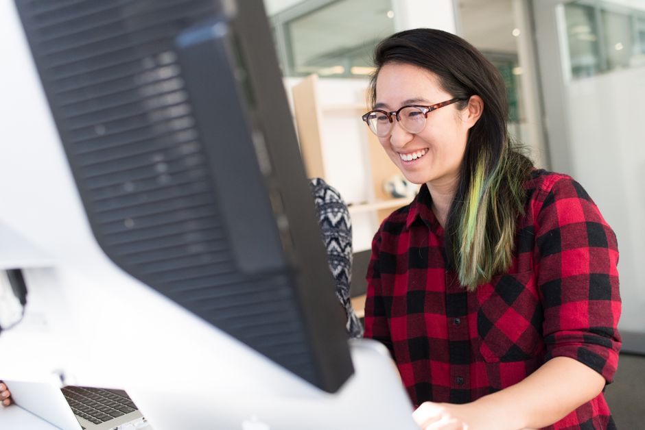 Smiling Asian woman with eyeglasses working at a desk in a modern office interior.