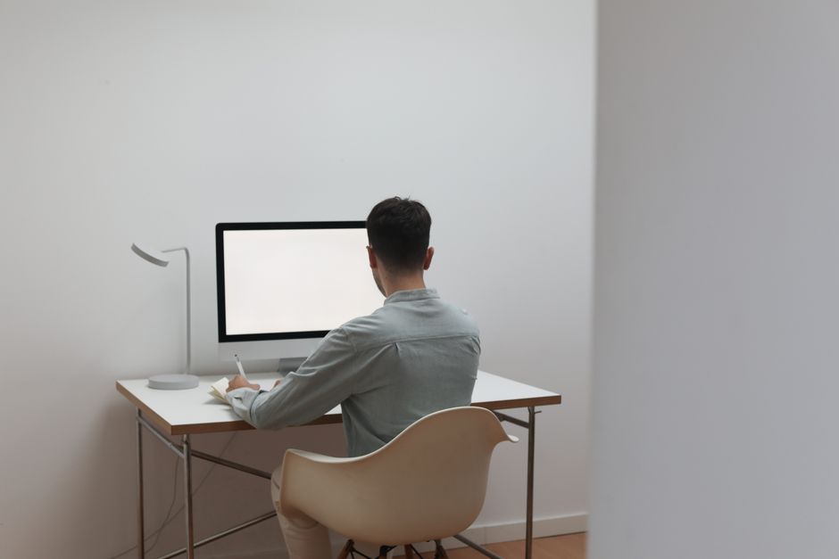 Back view of anonymous male entrepreneur sitting at table with computer and taking notes while working in light room