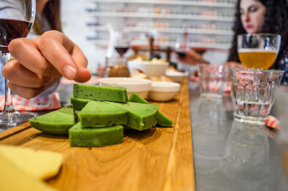 Woman reaching for colorful green cheese on a dining table indoors, surrounded by drinks and snacks.