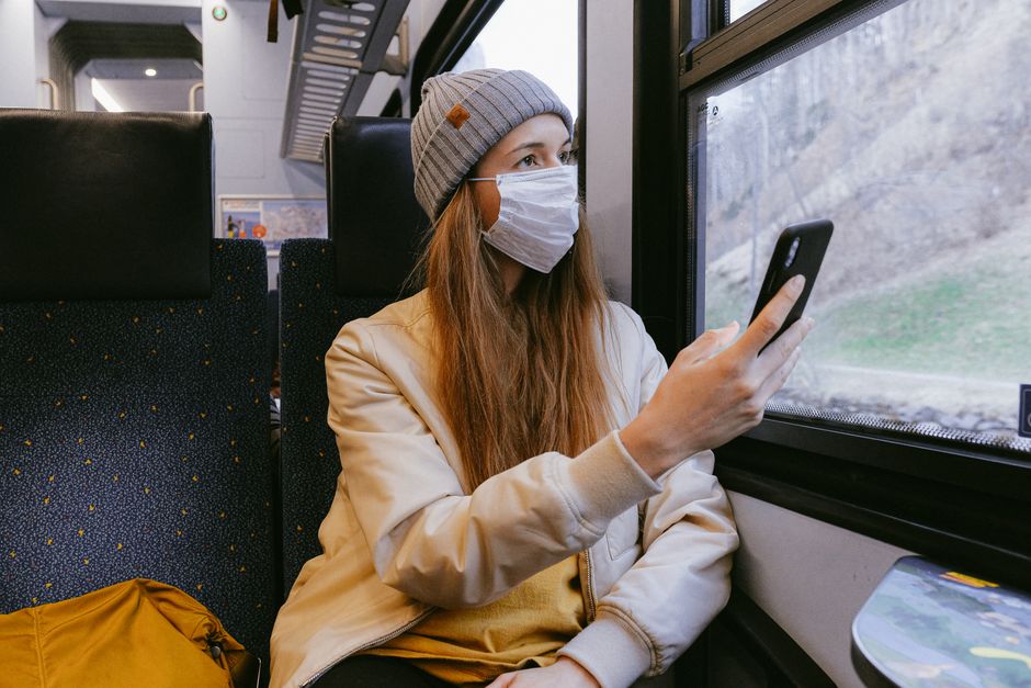Woman in casual wear wearing a face mask, using a smartphone on a train, reflecting pandemic lifestyle.