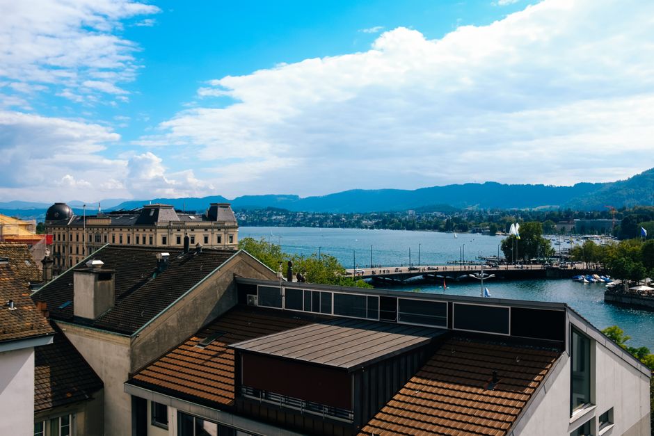 Panoramic view of Zurich featuring the Limmat River, rooftops, and mountains under a bright blue sky.