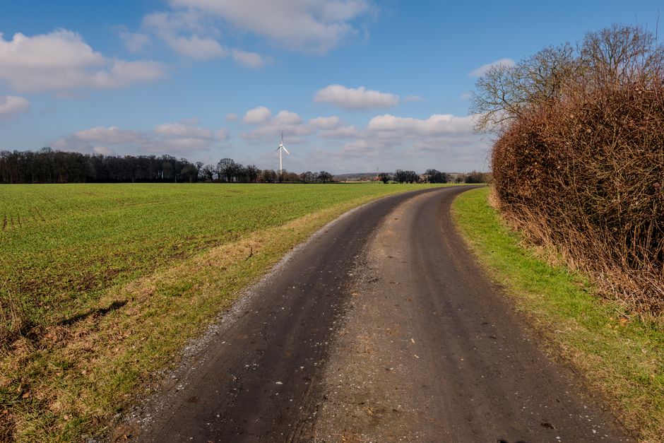 A scenic rural pathway with a distant wind turbine, surrounded by fields and a blue sky.