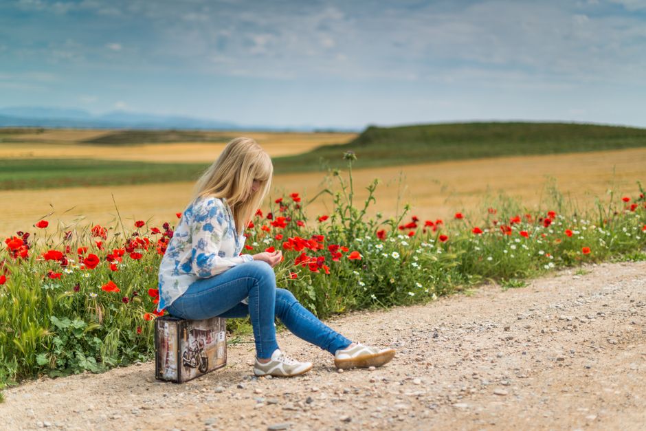 Blonde woman sitting on a suitcase by a dirt road surrounded by red poppies and green fields under a clear blue sky.