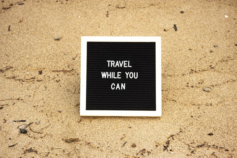 Quote 'Travel While You Can' on board in sand, inspiring travel and adventure.