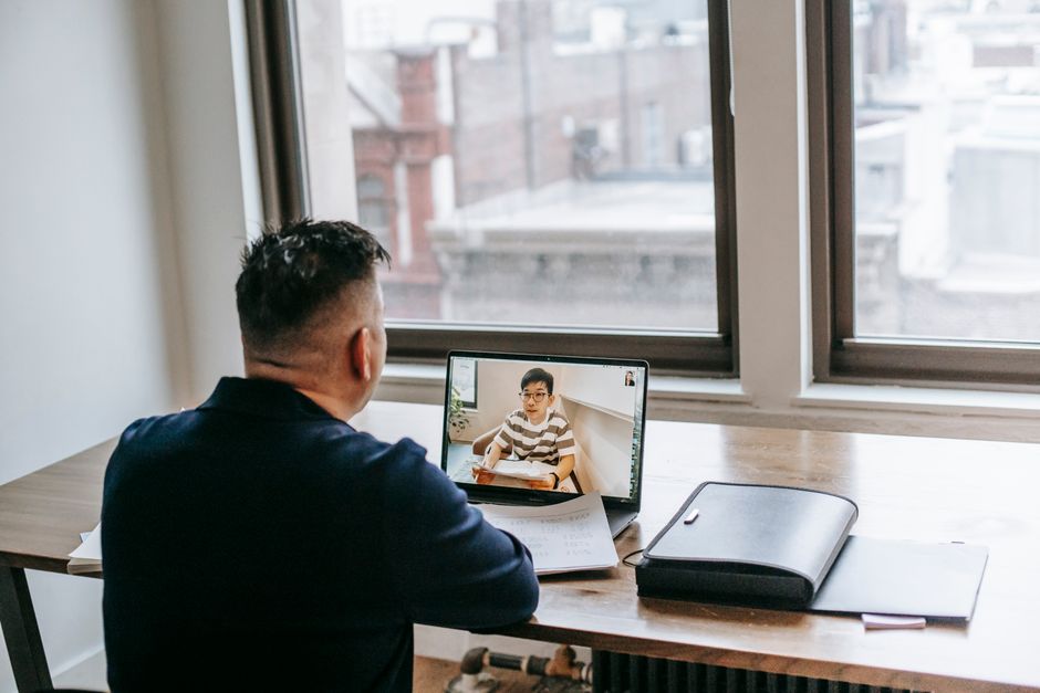 Back view of male in casual clothes sitting at table on chair while having video chat on laptop with pupil near papers and briefcase in light apartment near window